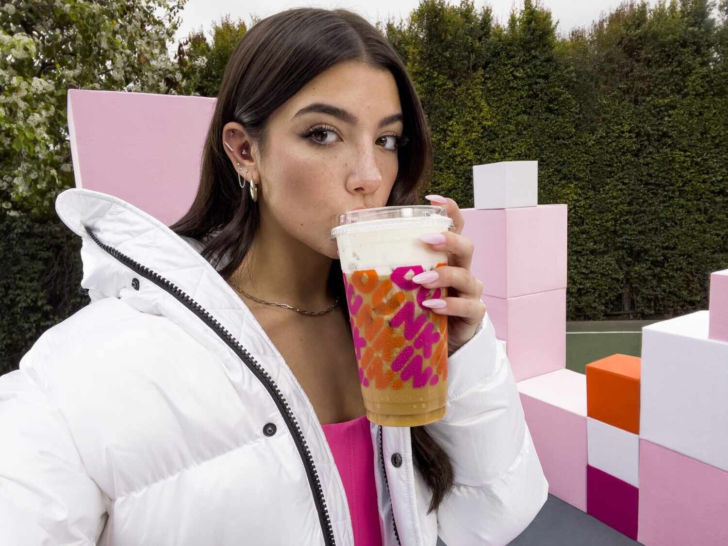 A young woman with long brown hair is pictured wearing a white winter coat and sipping from a large Dunkin’ Donuts iced coffee.